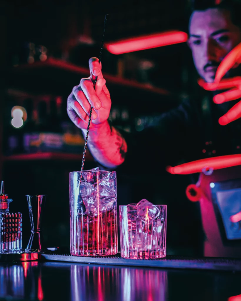 Un bartender concentra l'attenzione mentre miscela un cocktail con un lungo cucchiaio da bar, illuminato da luci colorate che creano un'atmosfera avvolgente.