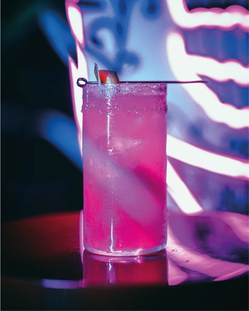 Un cocktail rosa in un bicchiere alto, guarnito con una fettina di agrume, brilla contro uno sfondo luminoso al neon.